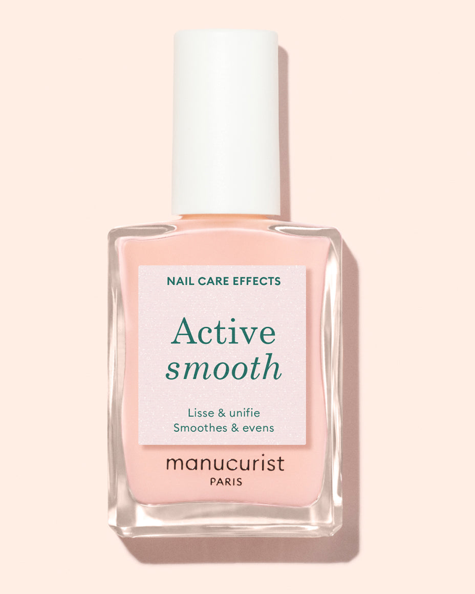 A bottle of manucurist Paris nail polish called Active Smooth with pale pink polish inside. The label highlights its nail care effects, stating it smoothes and evens nails. The background is a soft peach color.