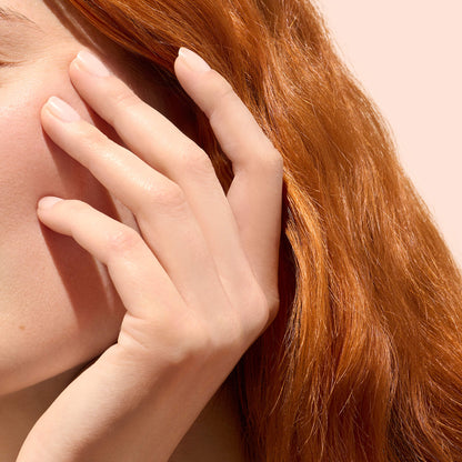 A close-up of a person with long red hair touching their face, showcasing their hand adorned in Manucurist’s Active™ Smooth 00 plant-based nails. Only part of the face, hand, and hair are visible against a soft, light background.