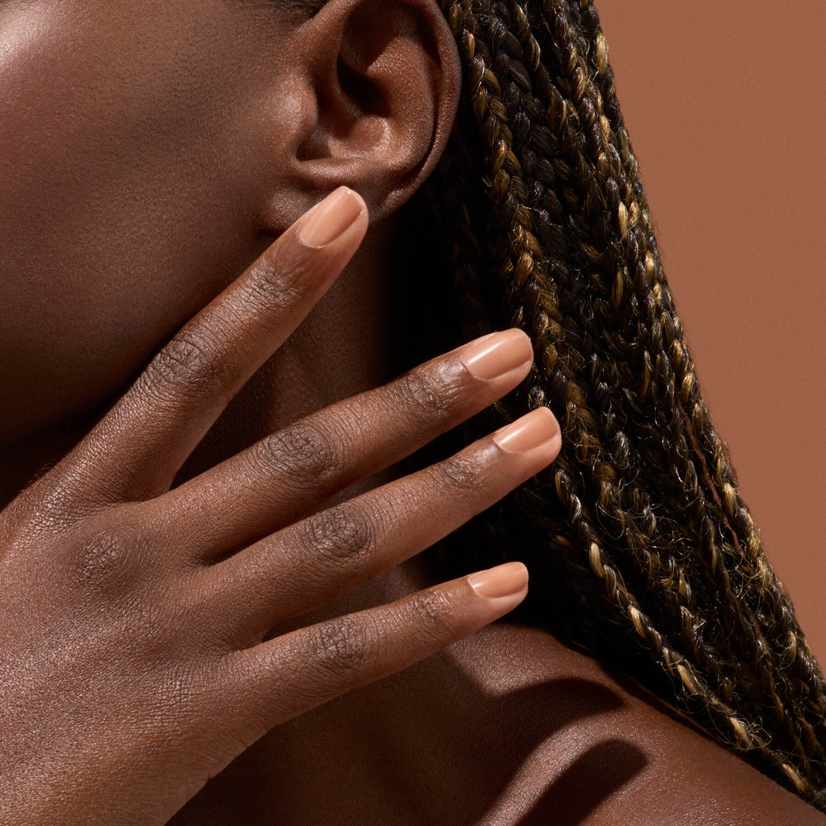 Close-up of a person with braided hair touching their face, showing neatly manicured fingernails painted in a nude color against a brown background.