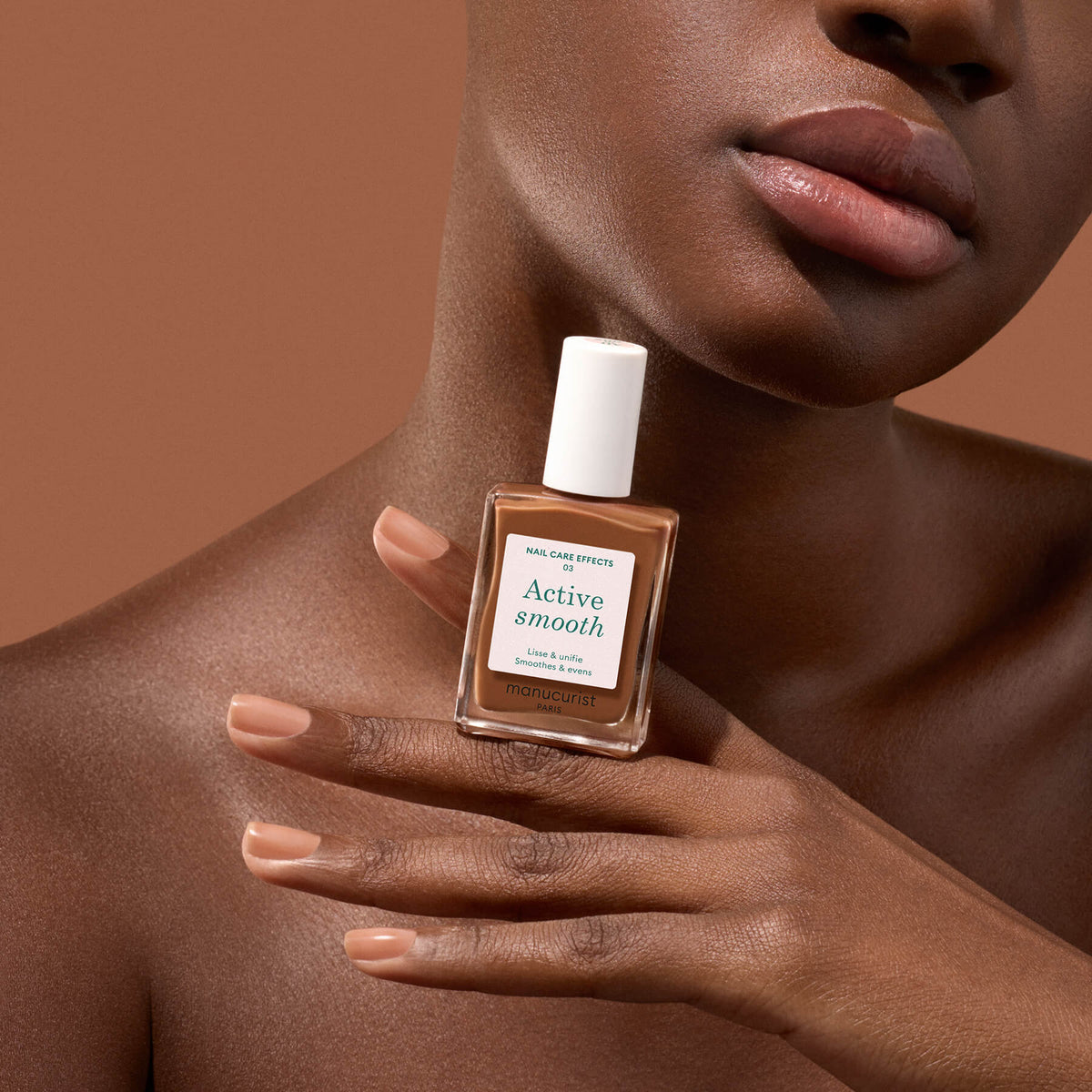 A person with smooth brown skin holds a Manucurist Active Smooth 03 nail polish near their face, displaying nude manicured nails against a matching brown background to showcase the benefits of quality nail care.