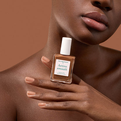 A person with smooth brown skin holds a Manucurist Active Smooth 03 nail polish near their face, displaying nude manicured nails against a matching brown background to showcase the benefits of quality nail care.