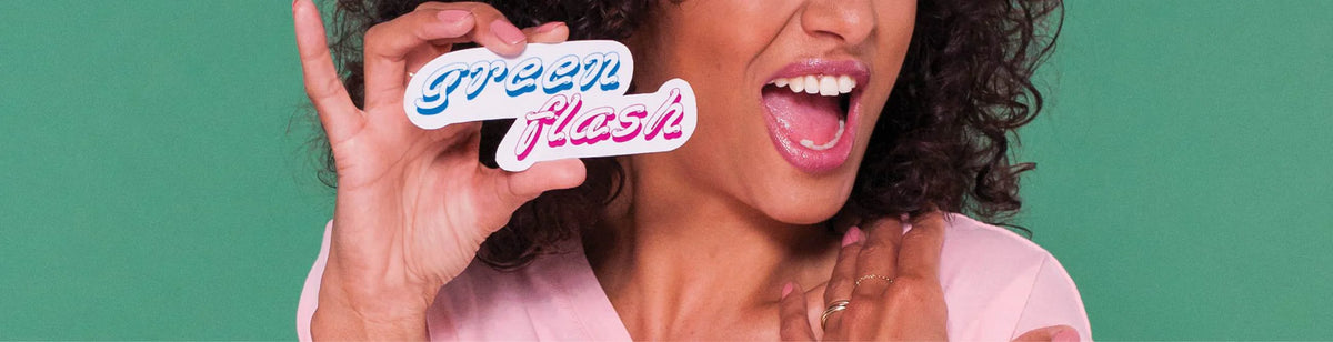 A smiling woman with curly hair holds a sticker reading green flash in colorful script, while wearing a light pink top, against a green background.