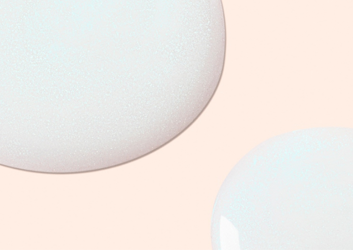 Two large, glossy, white liquid drops with smooth edges are set against a pale peach background. The drops appear slightly translucent and have a shiny surface that reflects light.