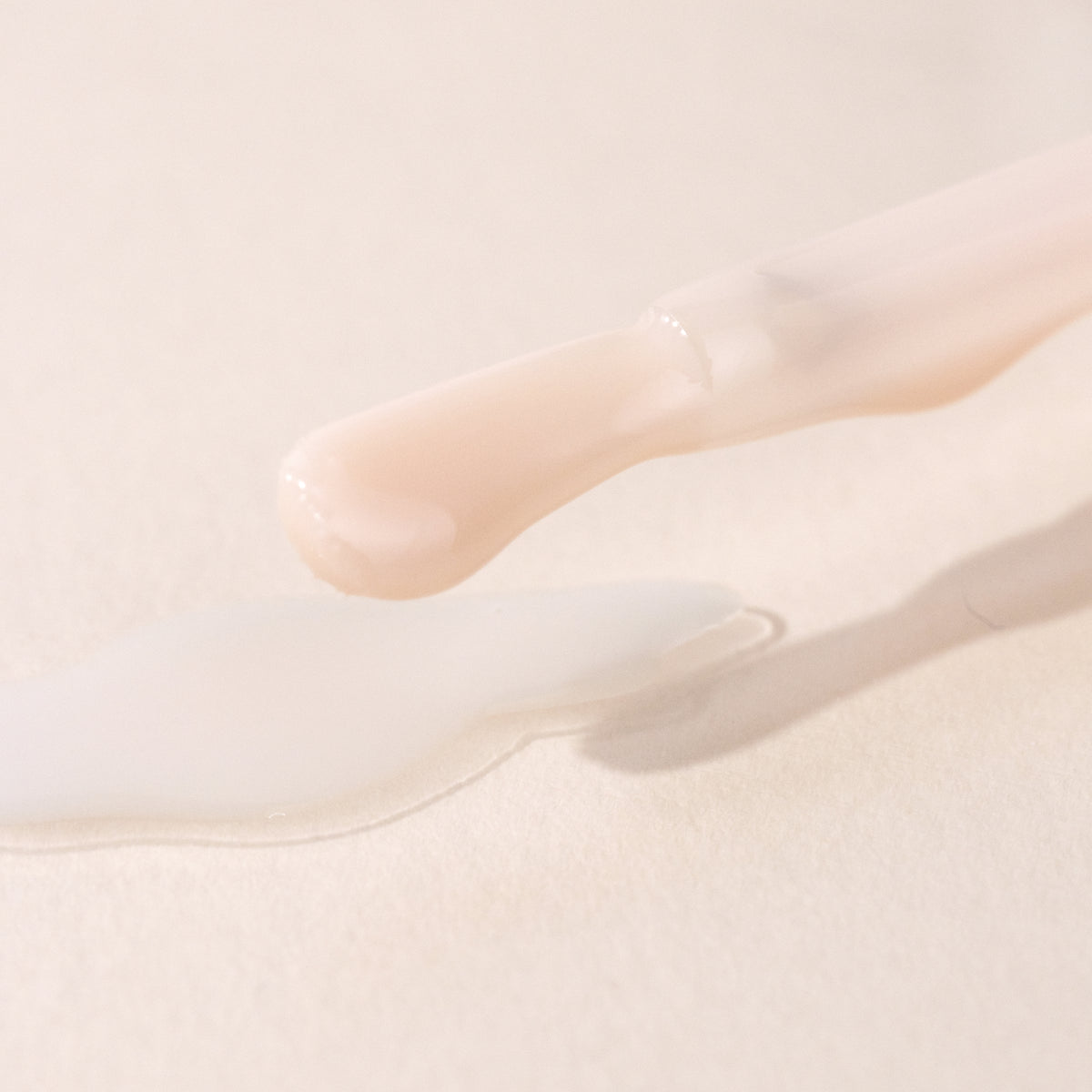 A close-up of a nail polish brush with pale pink polish, hovering above a spill of the same color on a light, textured surface.