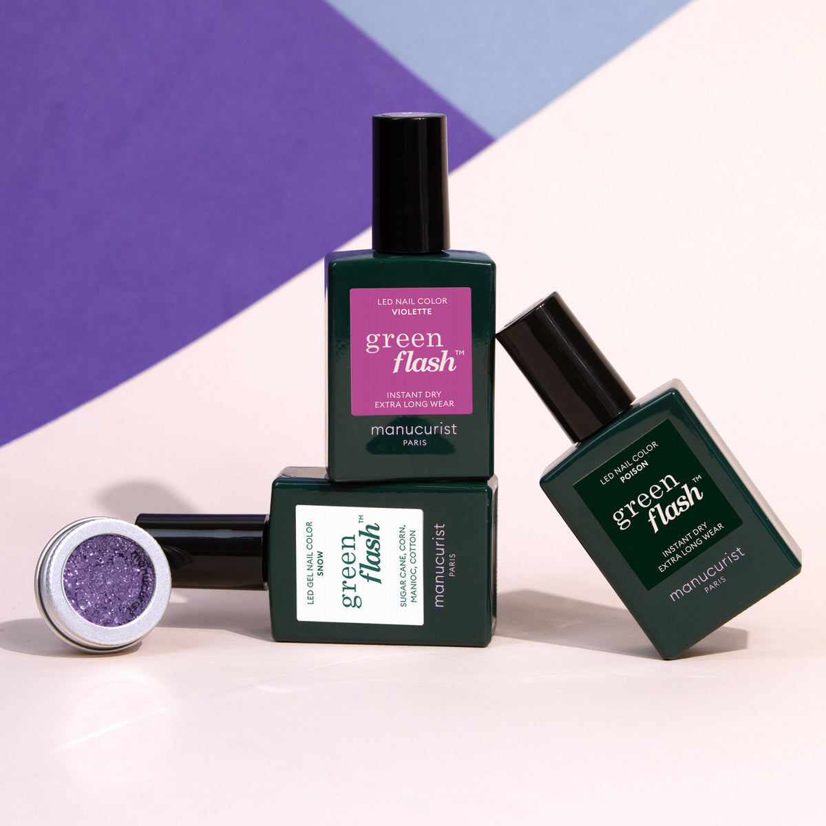 Three bottles of Manucurist Witchy  Wicked gel polish, stacked with one on its side, are shown next to a small pot of biodegradable glitter on a color-blocked background.