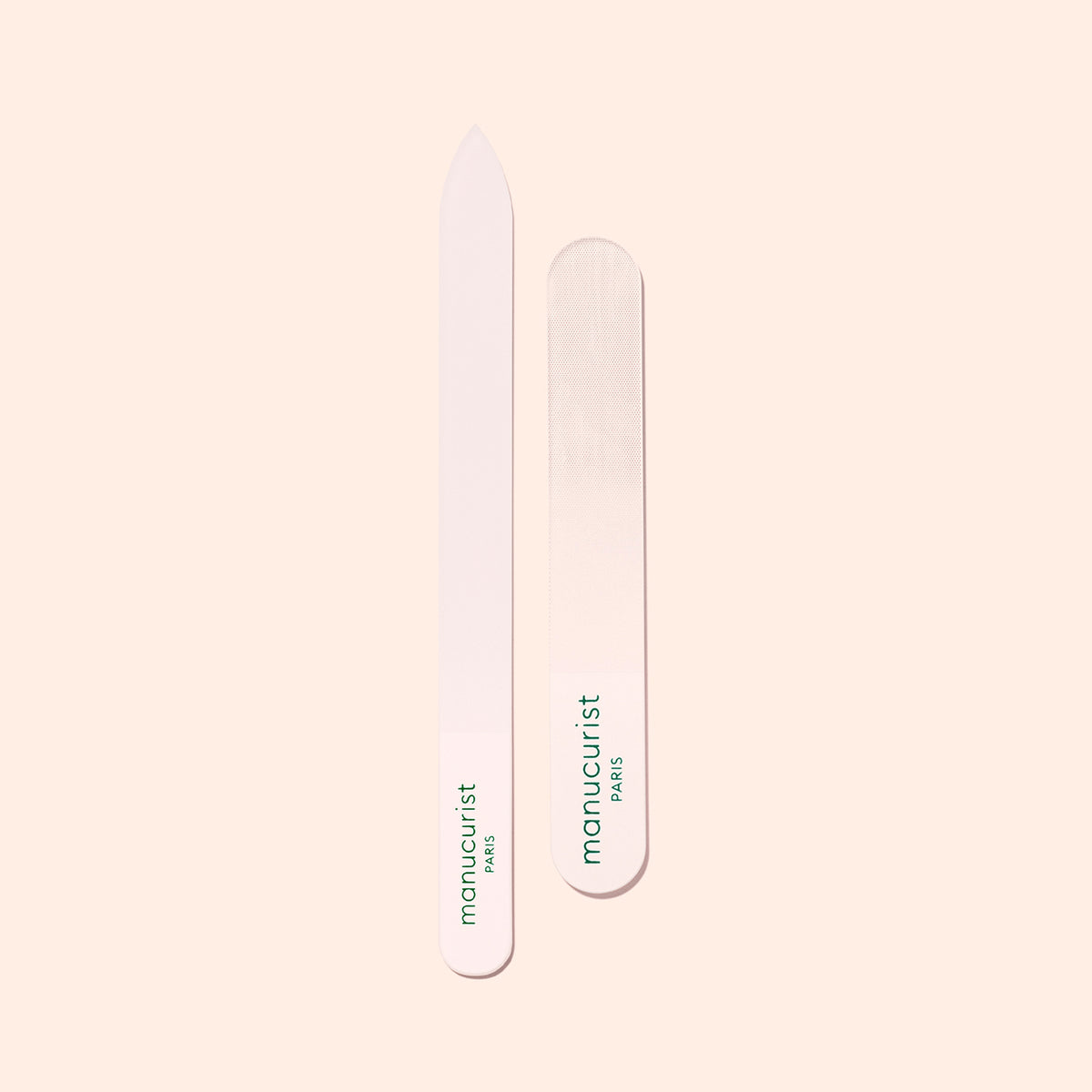 Two Manucurist-us Glass Nail Files, each with green "manucurist PARIS" text, are shown vertically on a light beige background. One file features a pointed end while the other has a rounded end.