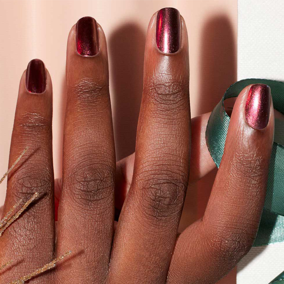 Une main à la peau foncée présente des ongles courts et soignés vernis avec la teinte Utopia de Manucurist — un vernis gel rouge cramoisi brillant — tout en tenant un ruban bleu canard sur un fond légèrement flouté.