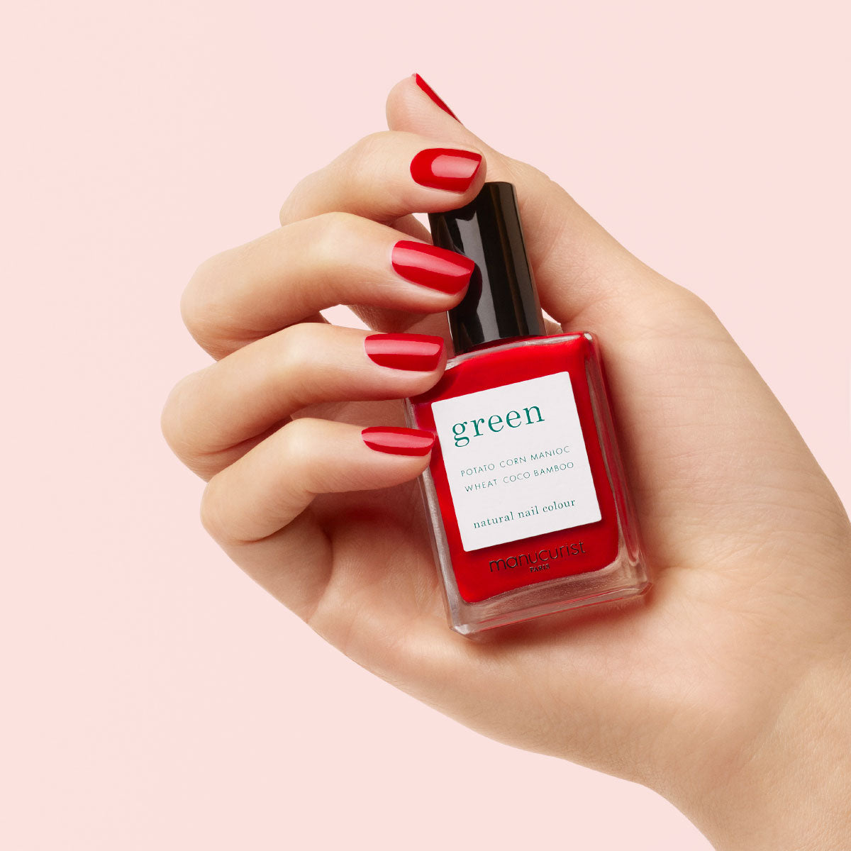 Une main aux ongles rouges soigneusement manucurés tient un flacon de vernis à ongles rouge étiqueté « green » par Manucurist, sur un fond rose clair.