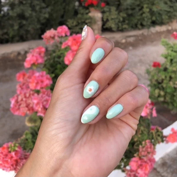Une main aux ongles longs et en amande, vernis en bleu pastel clair, avec un Nail Art floral blanc et orange sur un doigt, posée devant des fleurs roses et de la verdure en extérieur.