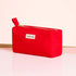 The Manucurist-us Red Travel Bag, a rectangular organic cotton pouch with a zipper and side handle, stands on a glossy surface. A white label with the brand name is visible. The background is light pink with a wooden accent.