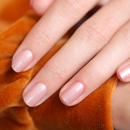 A close-up of a hand with neatly manicured nails painted in Carnation by Manucurist rests on orange velvet fabric.