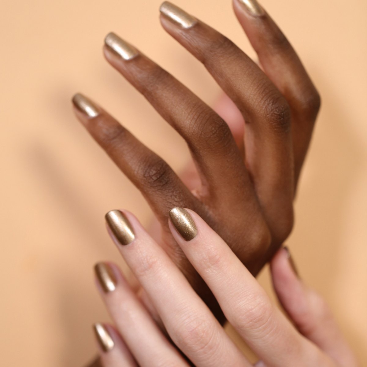 Two hands with varied skin tones, each sporting short, manicured nails painted in Manucurist’s Bronzé metallic gel polish, are beautifully posed against a soft beige background.