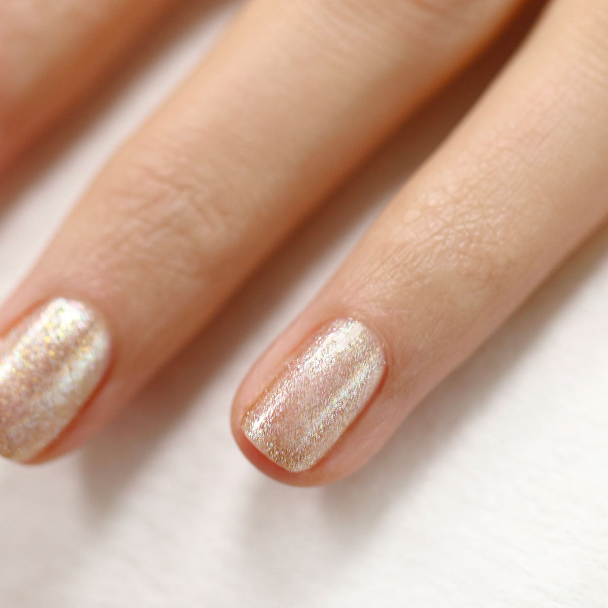 Gros plan sur deux doigts aux ongles courts et soignés, vernis avec la teinte Gold de Manucurist — un rose clair brillant et irisé rehaussé de paillettes dorées — sur fond blanc.