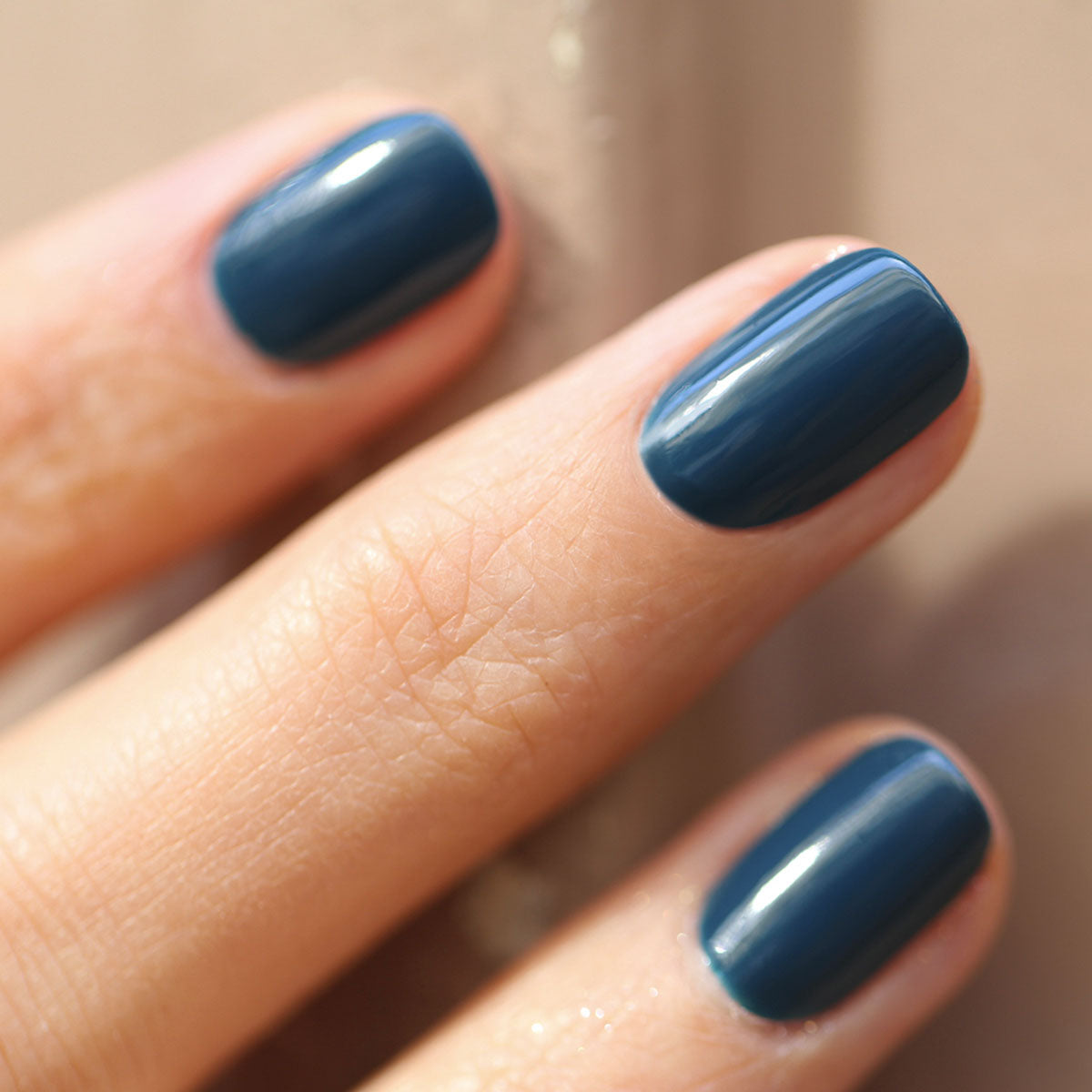 Gros plan d’une main aux ongles courts et soignés, vernis d’un bleu-vert brillant "Dark Clover" de Manucurist. Les doigts sont légèrement écartés sur un fond délicatement flou.