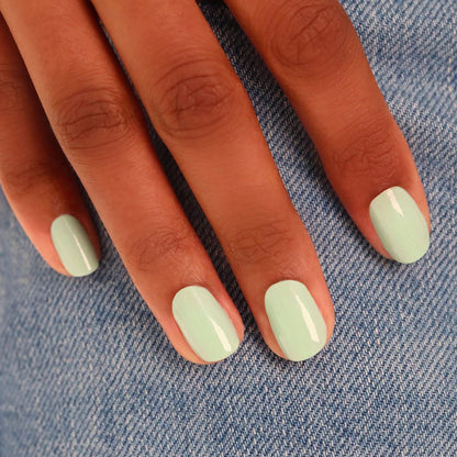 A hand with short, neatly manicured nails painted in Manucurist’s Mint, a glossy pastel green shade, rests on blue denim fabric.