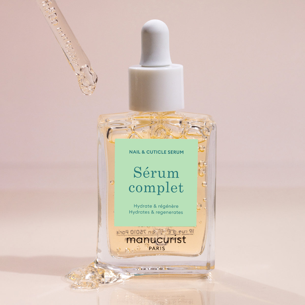A dropper releases Manucurist Nail Hydrating Serum into a clear glass bottle labeled with the brand name. The soft beige background highlights droplets of this nourishing nail treatment visible on the bottle.