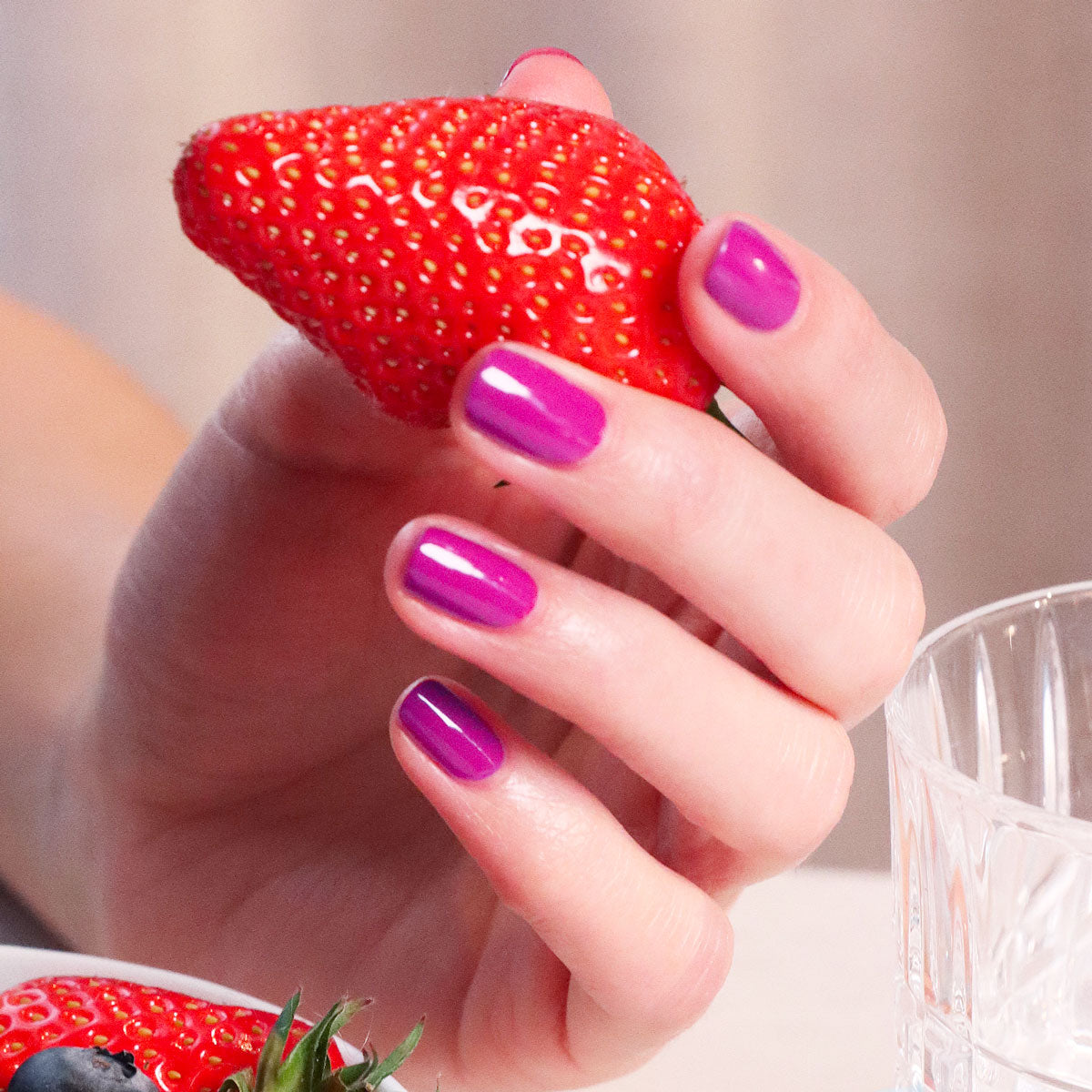 A hand with shiny, mauve Armeria gel polish by Manucurist holds a large red strawberry above a clear glass and some berries on a table.