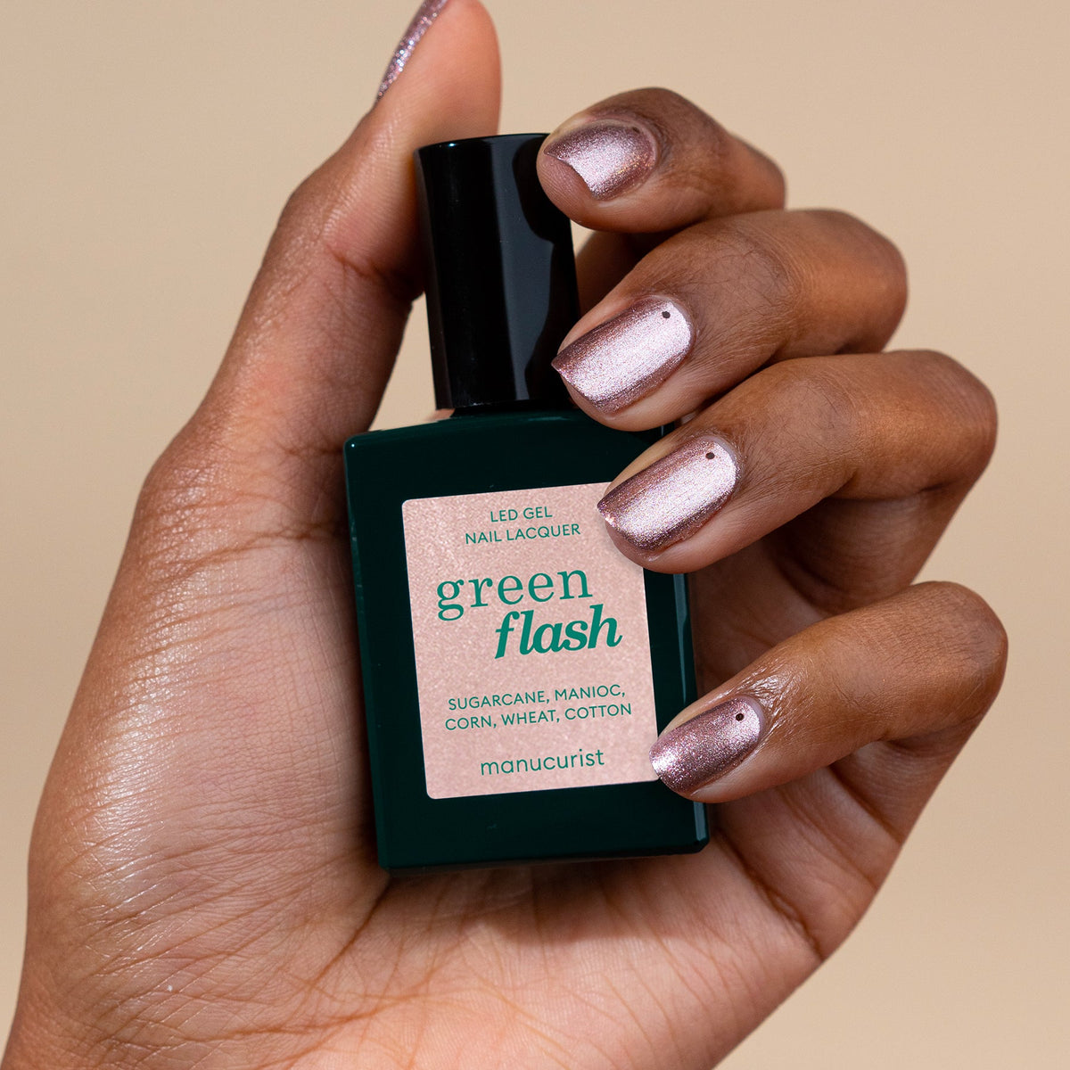 Une main au vernis gel nacré rose irisé tient un flacon de vernis gel Manucurist Cosmic Rose sur fond beige.