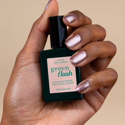 Une main au vernis gel nacré rose irisé tient un flacon de vernis gel Manucurist Cosmic Rose sur fond beige.