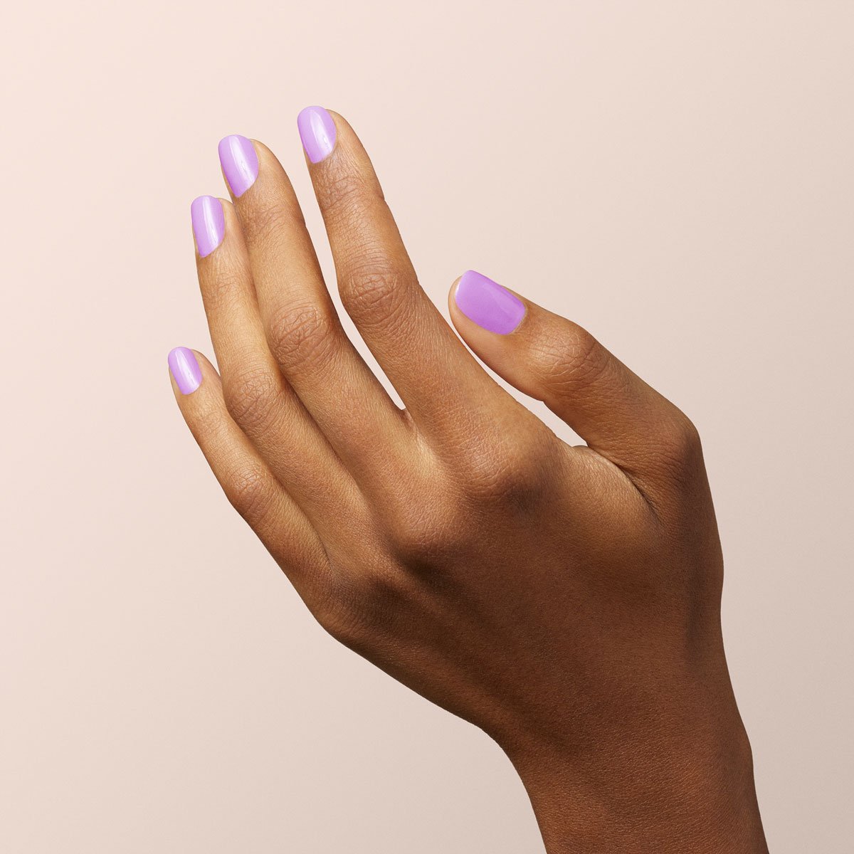 A hand with medium brown skin is shown against a light beige background. The nails are manicured and painted with Lisa Lilas, a glossy lilac nail polish from Manucurist.