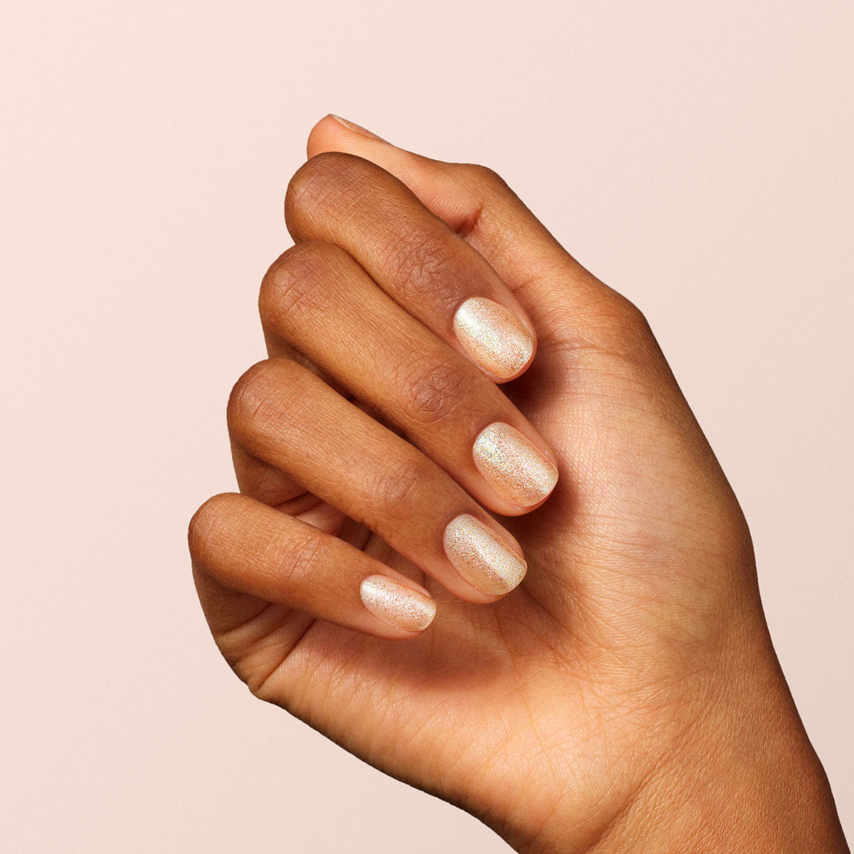Une main à la peau mate présente des ongles courts et soigneusement manucurés, vernis avec "Gold" de Manucurist, un vernis pailleté pêche clair scintillant, sur fond beige doux.