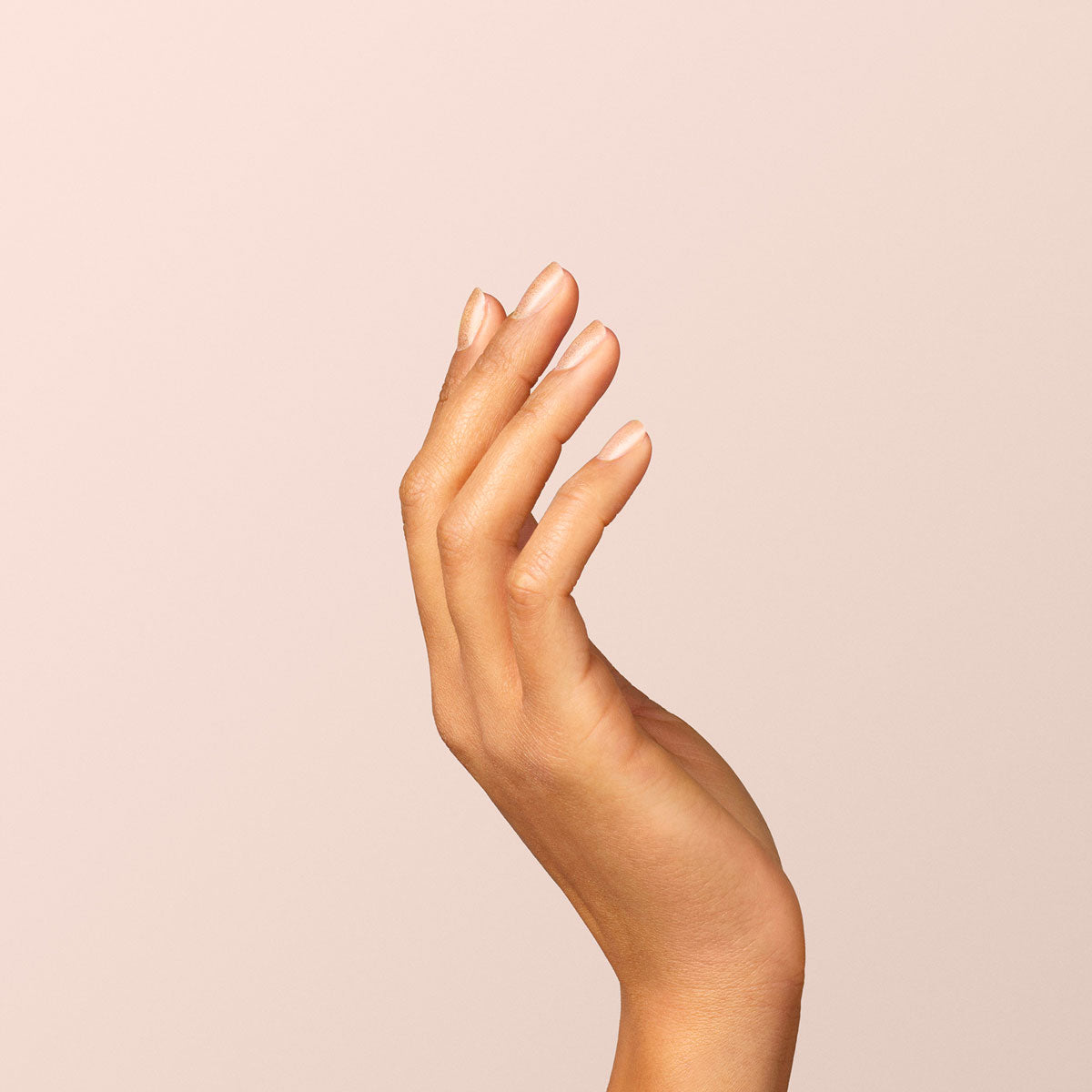 Une main aux ongles soigneusement manucurés, certains vernis avec la teinte Gold de Manucurist, est levée sur un fond beige clair uni. Les doigts sont légèrement courbés et détendus, mettant en valeur une peau lisse et des ongles élégants et naturels.