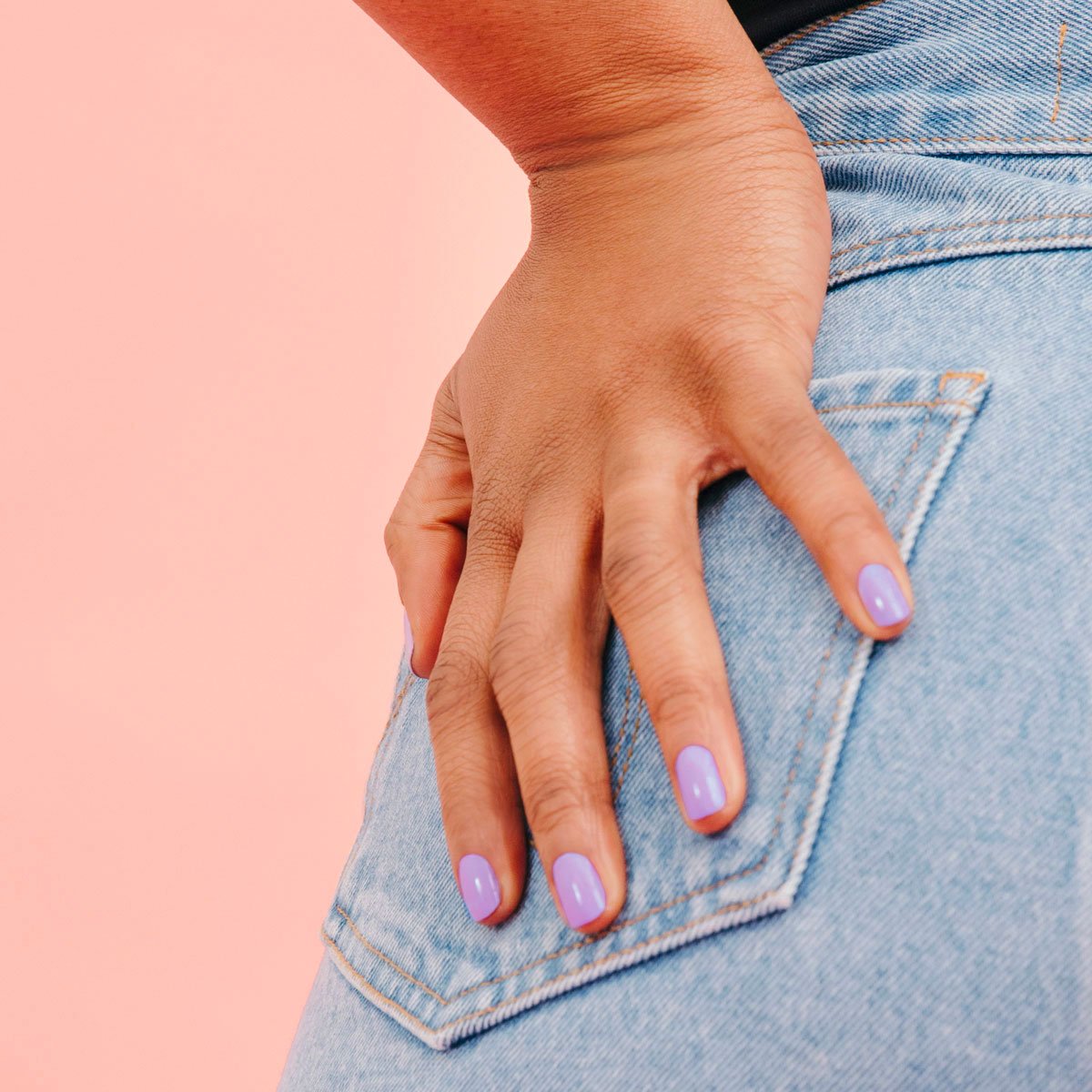 Une main avec le vernis gel Lisa Lilas de Manucurist repose sur la poche arrière d’un jean en denim bleu clair, sur fond rose pour une parfaite ambiance années 90.