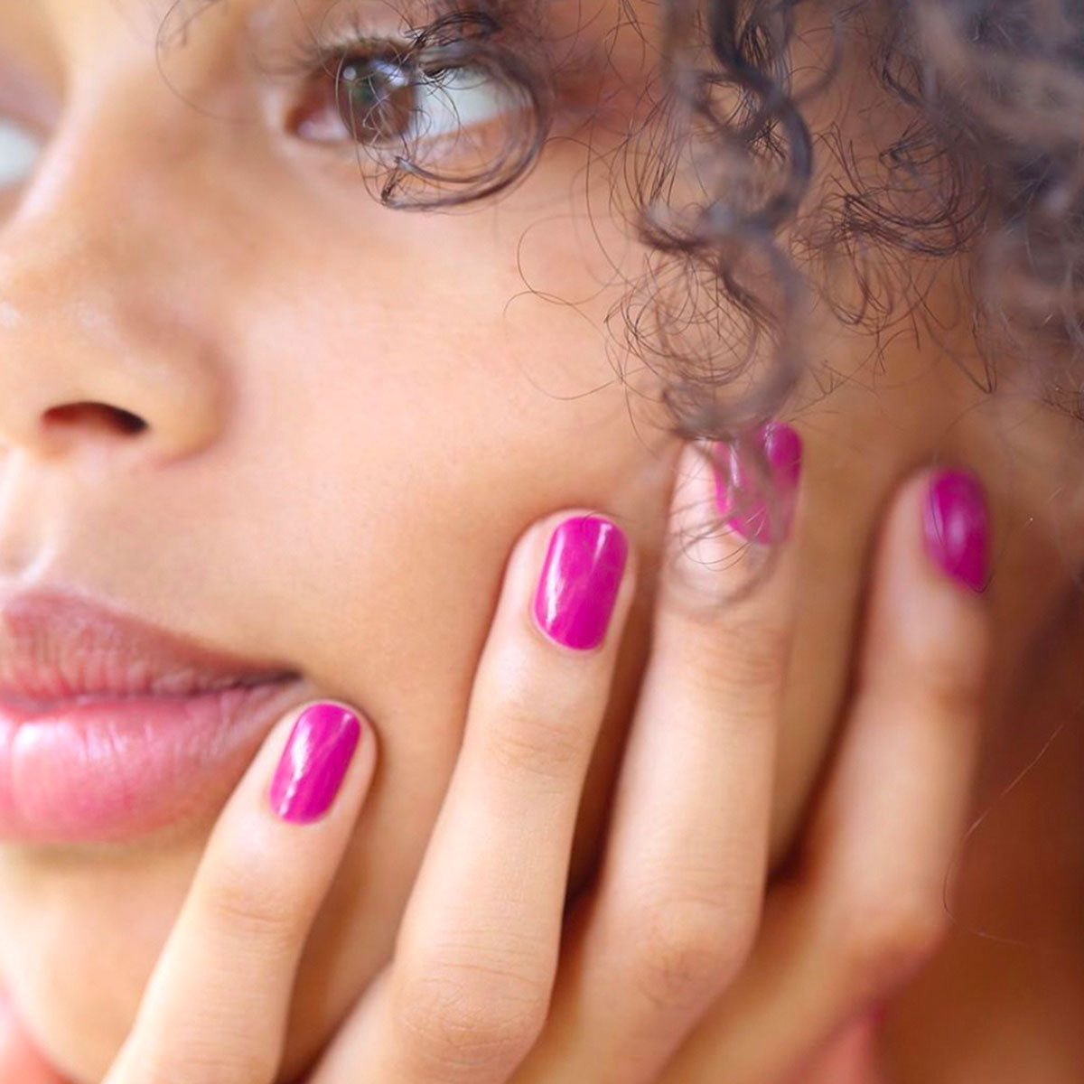 A close-up of a person with curly hair touching their cheek, showcasing glossy mauve "Armeria" nail polish from Manucurist&