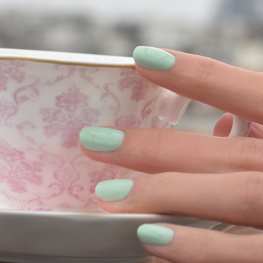 A hand with Manucurist Mint pastel green nail polish holds a white teacup with a pink floral pattern and gold rim against a blurred background.