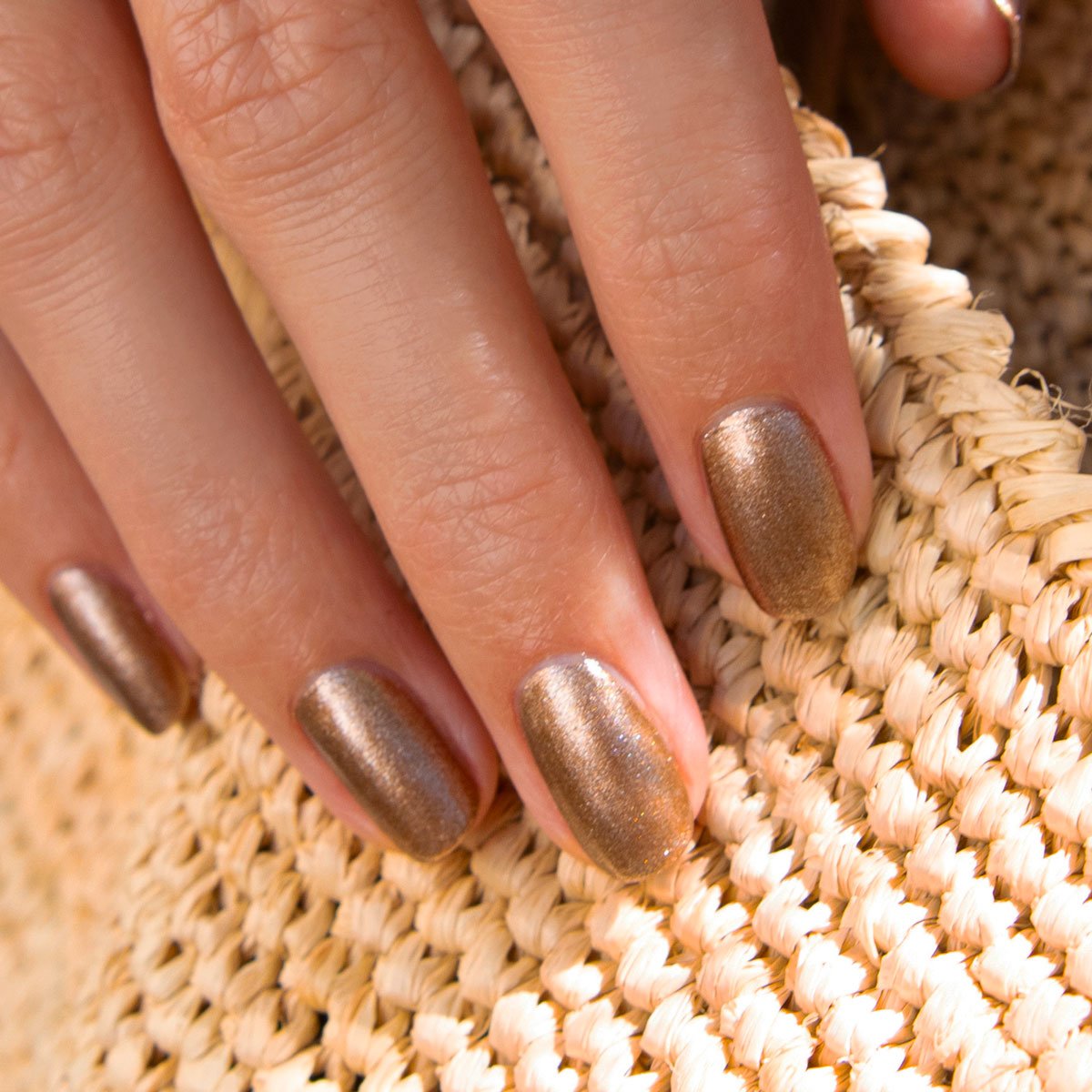 A close-up of a hand with short, neatly manicured nails painted in Manucurist’s Bronzé gel polish, resting on a woven straw surface.