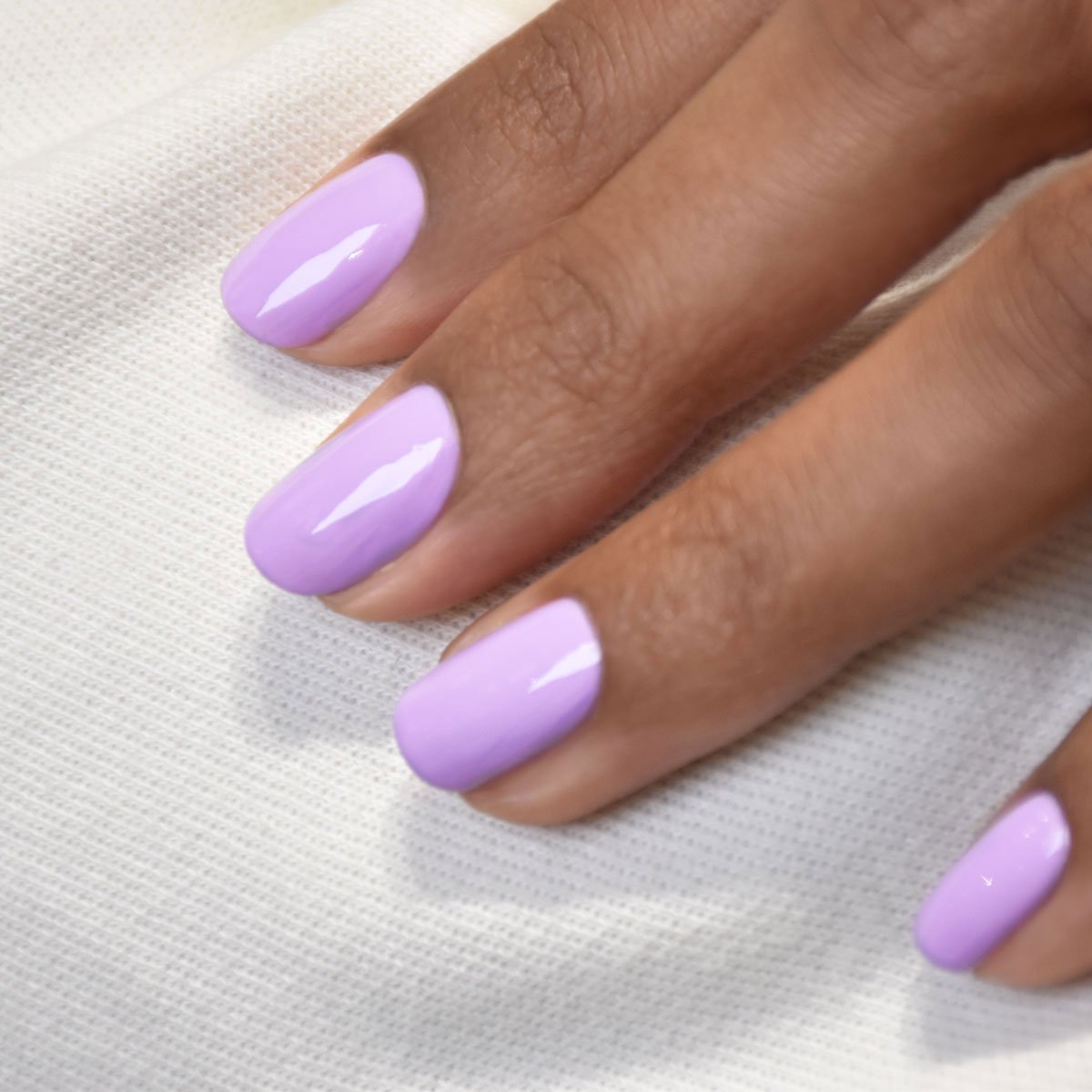 Une main aux ongles soigneusement manucurés, vernis avec le vernis gel Lisa Lilas de Manucurist, repose sur un tissu blanc texturé, évoquant de subtiles vibes des années 90.
