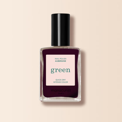 A bottle of Manucurist Aubergine nail polish, with a deep purple shade and a black cap, stands against a light beige background.