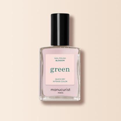 A bottle of Manucurist nail polish in the shade “Blossom,” featuring a creamy light pink color, black cap, and square transparent glass bottle from the Green range is displayed against a beige background.