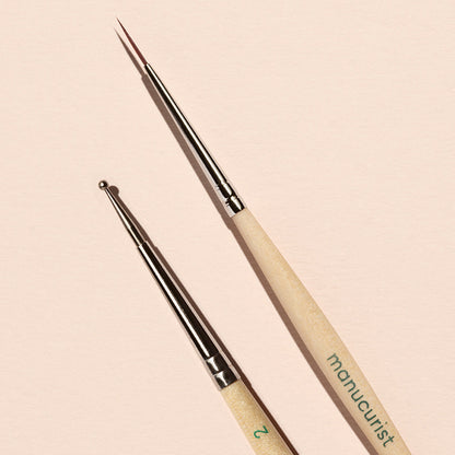The Manucurist Dotting Tool  Nail Art Liner Brush Duo features light wooden handles and silver tips, and is shown arranged on a pale pink background.
