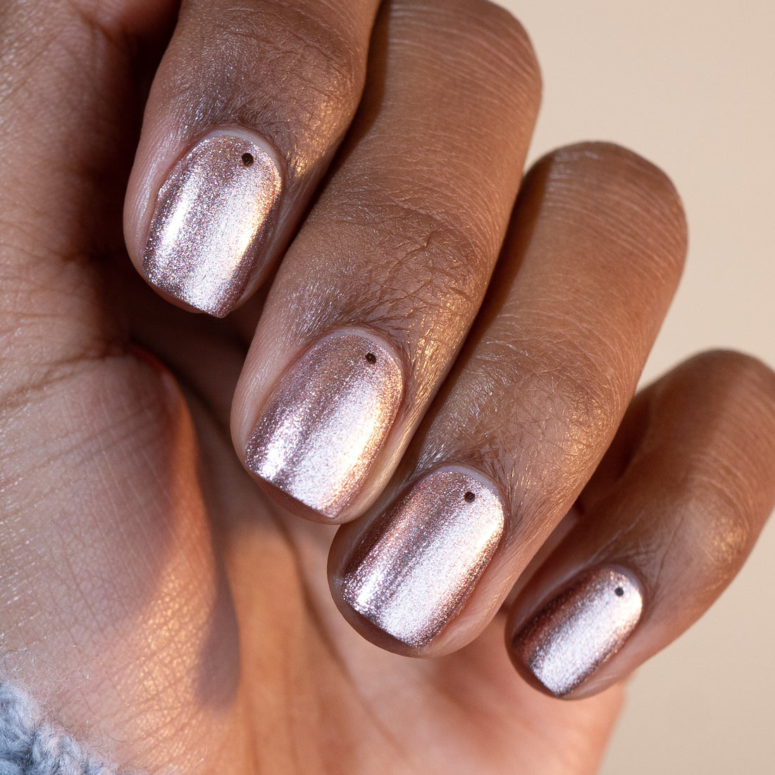 Une main avec le vernis Cosmic Rose de Manucurist — des teintes rose gold métallisé et rose irisé, chaque ongle surmonté d’un point noir près de la base — présentée sur un fond neutre.