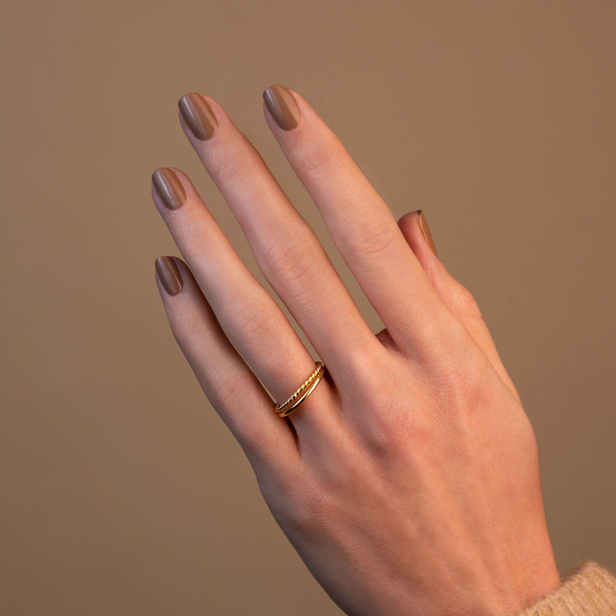 A hand with neatly manicured nails painted in Manucurist’s Orme polish, adorned with two delicate gold rings on the middle finger, is shown against a plain beige background.