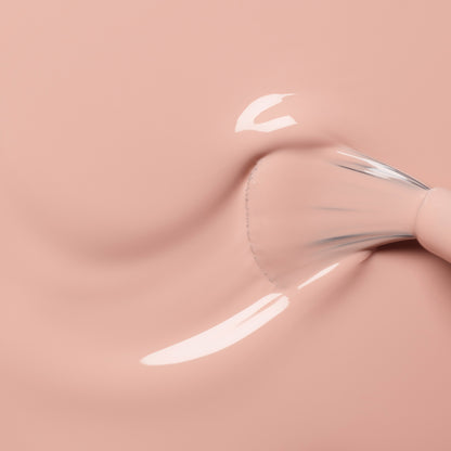 A close-up of a brush swirling Manucurist Dune foundation, a creamy light pink shade with orange undertones, on a smooth surface, creating a glossy texture and softly blurred streaks.