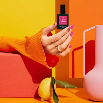 A hand with Pétula by Manucurist Green Flash gel polish holds a green nail polish bottle against an orange-yellow background, with a lemon and pink pitcher nearby. The hand, wearing an orange sleeve and white ring, rests on a red block.