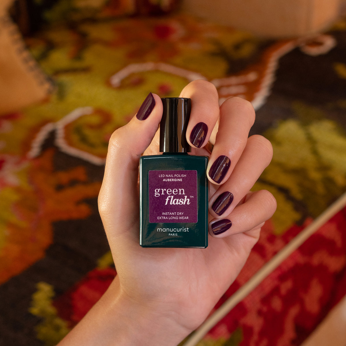 A hand with dark purple gel polish holds a bottle of Manucurist Aubergine nail polish against a blurred, colorful patterned background.