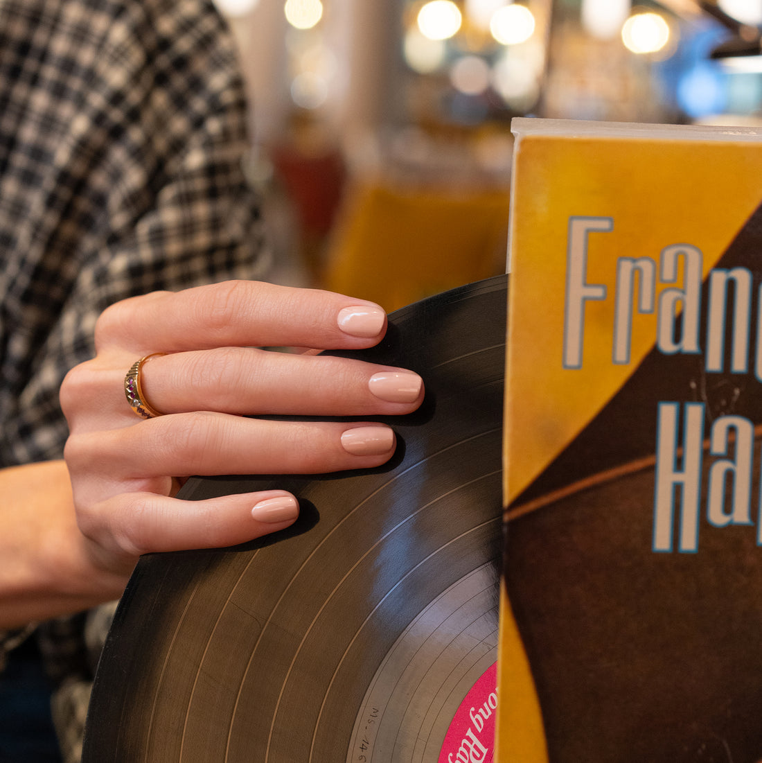 A hand with neatly manicured nails painted in Manucurist’s Dune shade holds a vinyl record beside a yellow album cover reading “Fran,” set against a softly blurred background with warm lights.