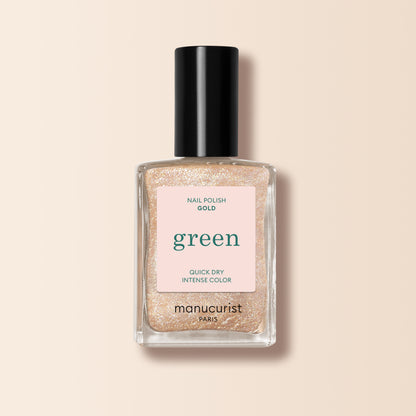 Un flacon de vernis à ongles Manucurist Gold de la gamme Green, avec un bouchon noir et une étiquette rose indiquant "green," "nail polish gold," et "quick dry intense color," présenté sur un fond beige.
