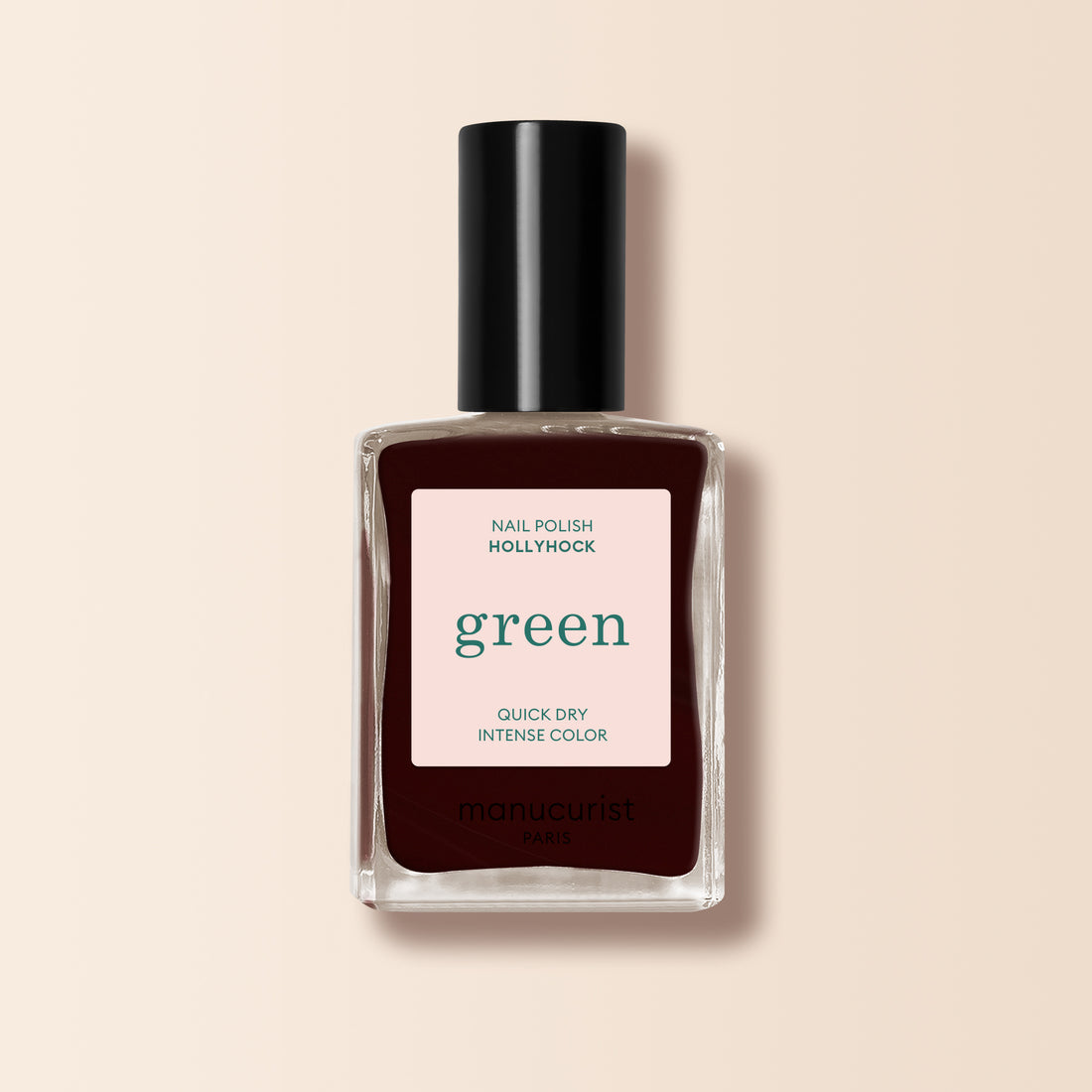 Un flacon de vernis à ongles Manucurist Hollyhock bordeaux profond, avec un bouchon noir et une étiquette indiquant "green, séchage rapide, couleur intense", est posé sur un fond beige.