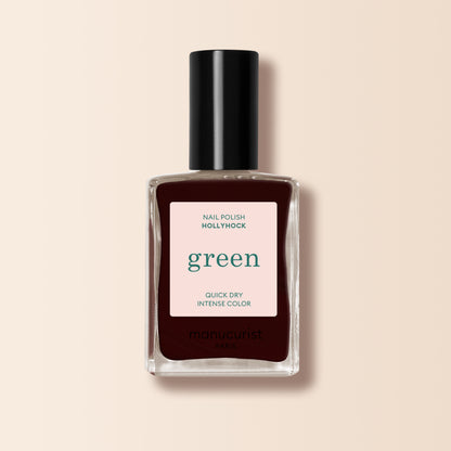 A bottle of Manucurist Hollyhock nail polish in deep burgundy with a black cap and a label stating "green, quick dry intense color" sits against a beige background.