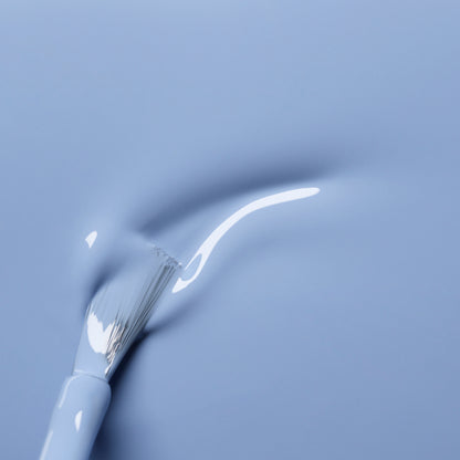 Gros plan sur le pinceau blanc Manucurist Lilas appliquant un vernis à ongles bleu pastel, traçant une ligne lisse et brillante sur une surface bleue assortie.