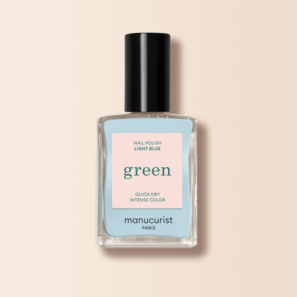 Un flacon de vernis à ongles Manucurist Light Blue de la gamme Green™, avec un bouchon noir et une étiquette « green quick dry intense color », est présenté sur un fond beige.