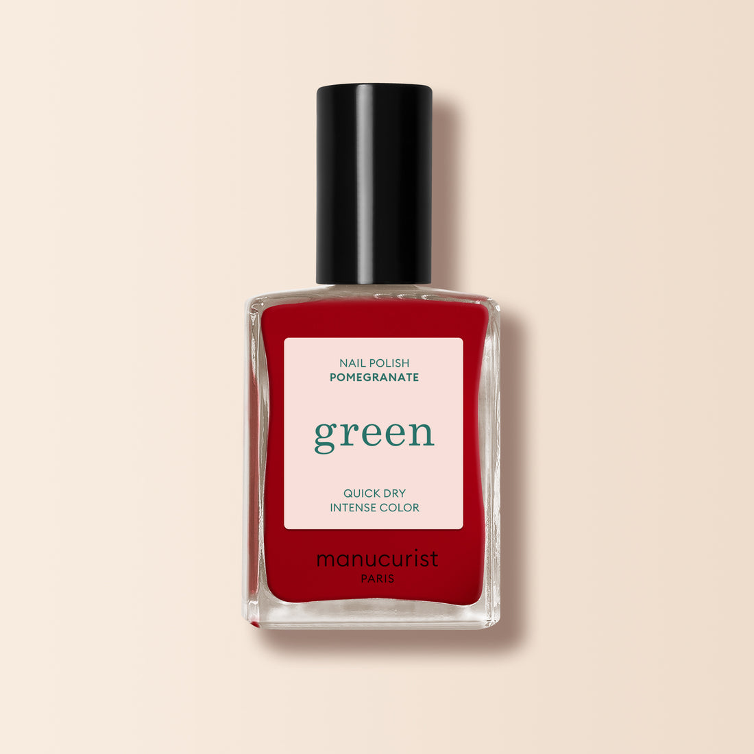 Un flacon de vernis à ongles Manucurist Pomegranate, dans une teinte rouge avec un bouchon noir et une étiquette blanche affichant du texte vert et rouge, est présenté sur un fond beige clair.