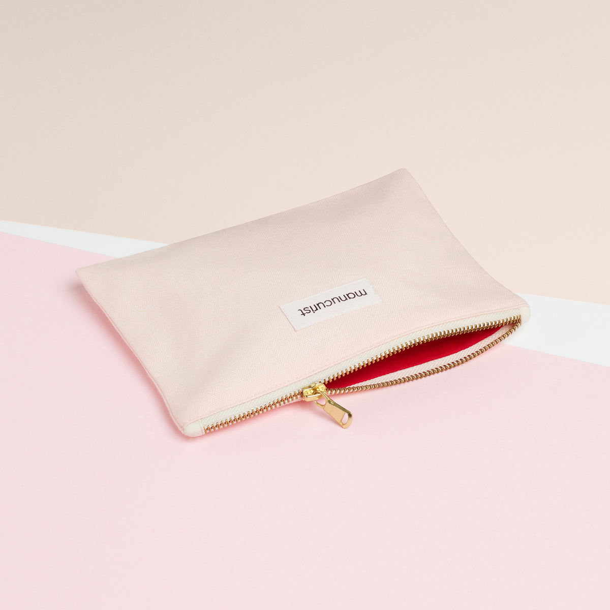 La Petite Trousse Rose Manucurist-us, en coton biologique avec une fermeture éclair dorée et une étiquette blanche « momentist », repose sur un fond rose et crème. Son intérieur doublé de rouge est parfait pour ranger vos essentiels beauté.
