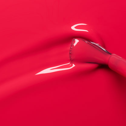 A close-up of a makeup brush spreading Manucurist Bougainvillea, a bright reddish-pink nail polish with glossy reflections on the smooth surface.