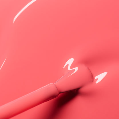 Close-up of a brush applying Manucurist Capucine, a glossy coral pink nail polish with a smooth, shiny finish and visible reflections.
