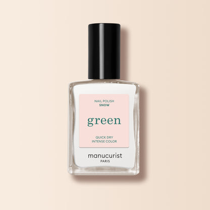 A clear glass bottle of Manucurist Snow nail polish with a black cap, labeled "Green." The label reads "Nail Polish Snow," offering pure white, quick-dry intense color, displayed on a light pink background.